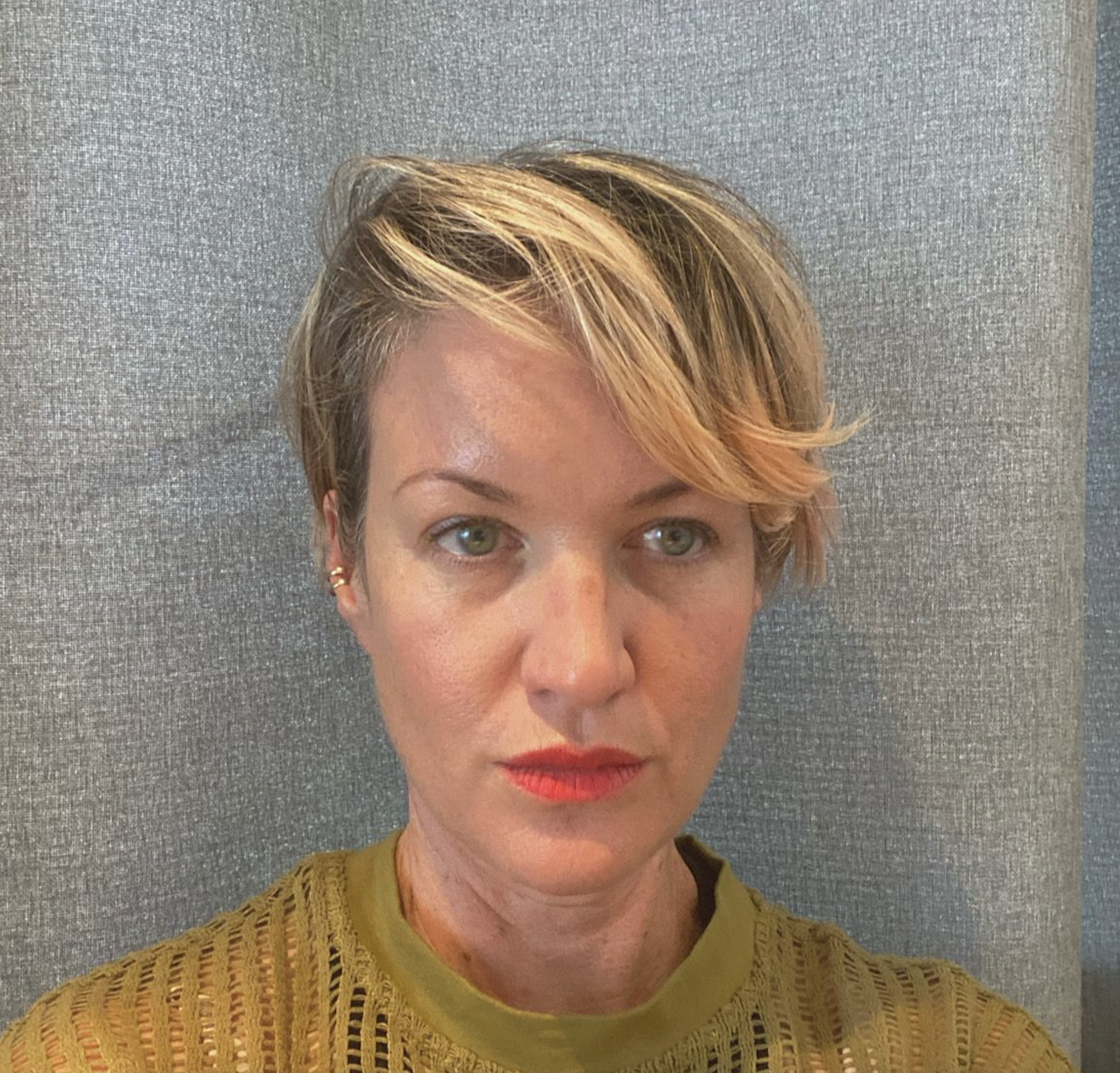A caucasian woman with short blond hair, green eyes, and a yellow top in front of a grey background